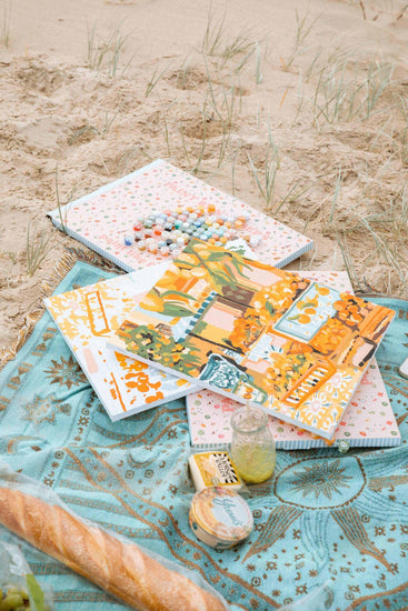 paint by numbers kits sitting on a picnic blanket on a beach